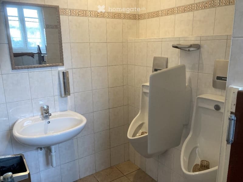 Public restroom with white tiled walls, a sink, two urinals, a mirror, and soap and paper towel dispensers.