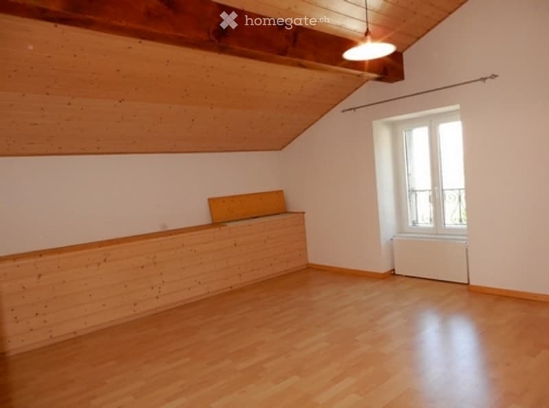 Empty room with wooden flooring, white walls, and a slanted wooden ceiling. A small wooden bench is placed against one wall, and a window with white frames is visible on the opposite wall.