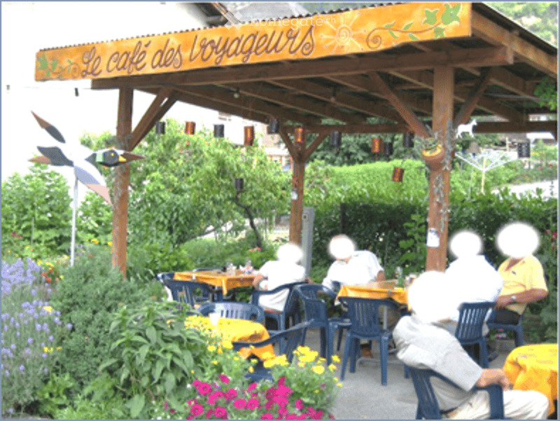 Outdoor seating area of a café with tables, chairs, and colorful flowers, under a wooden canopy with a sign reading "Le café des Voyageurs".