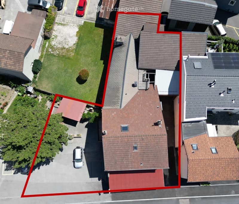 Aerial view of a multi-story residential building with a red-tiled roof, surrounded by neighboring structures and a small garden area.