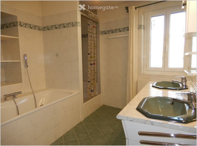 Bathroom with a bathtub, double sink, and a shower area with a curtain. The walls are tiled with a decorative border, and the floor is green tile.