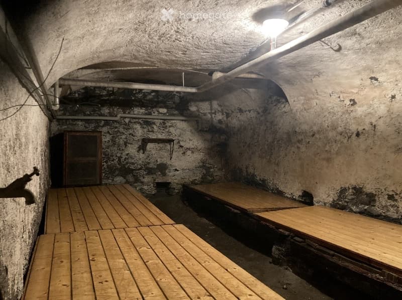 Underground room with stone walls, wooden benches, and a small wooden cabinet. Exposed pipes and a single light fixture are visible.