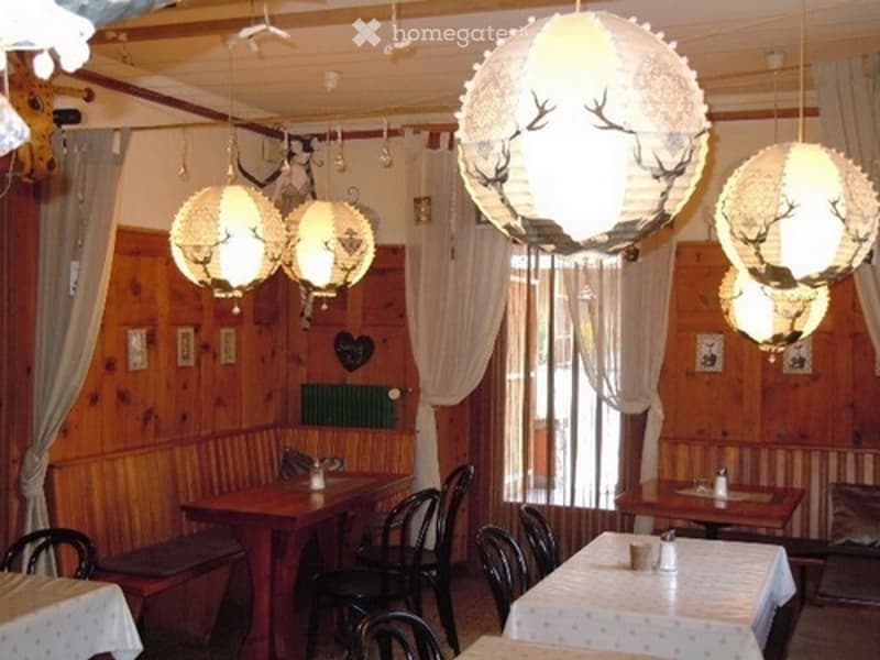 A cozy restaurant interior with wooden walls, large windows with curtains, and several tables and chairs arranged for dining. The space features decorative hanging lamps with a deer motif.