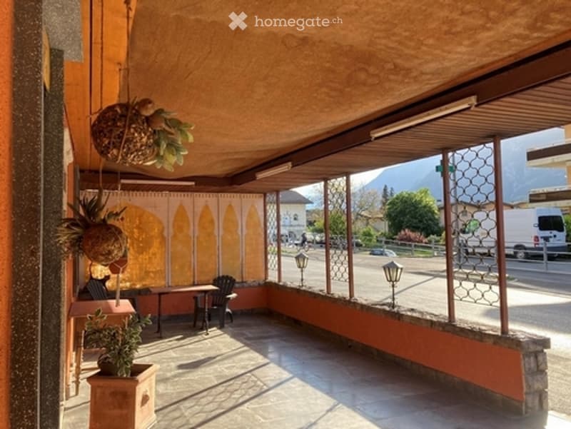 Balcony with wooden ceiling, orange walls, hanging plants, small table and chairs, outdoor lighting, overlooking a street with mountains in the background.