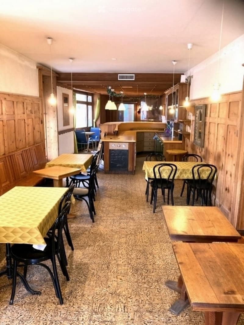 Restaurant interior with wooden tables, chairs, and benches arranged around a central area, with a kitchen counter visible in the background.