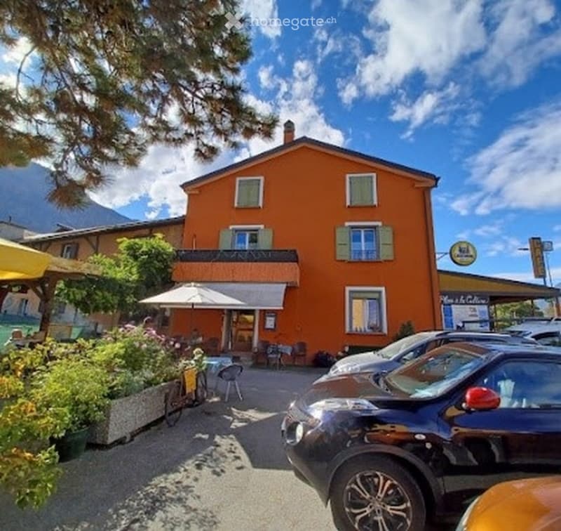 A two-story orange building with green shutters, located in a scenic area with mountains in the background. The building has a balcony and a small outdoor seating area with tables and chairs.