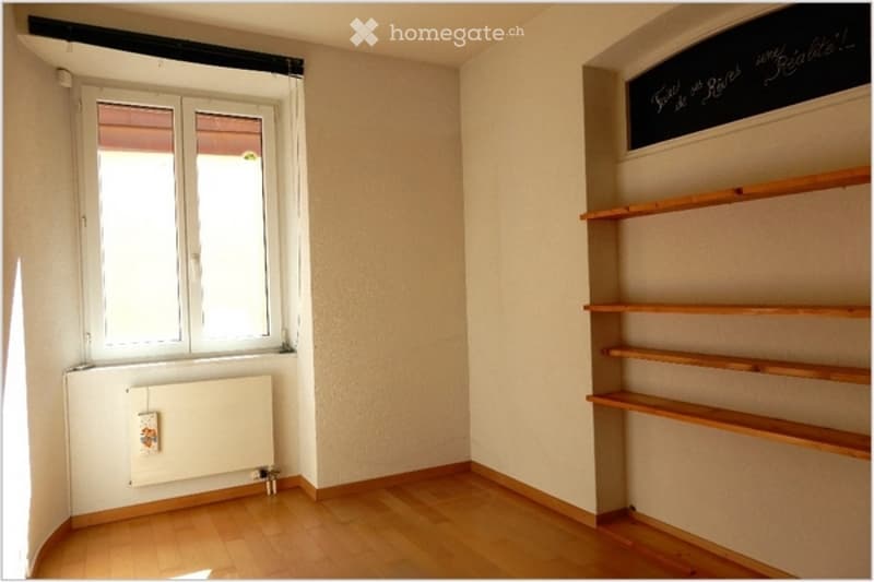 Empty room with wooden flooring, white walls, a window with white frame, a radiator, and wooden shelves on one wall.
