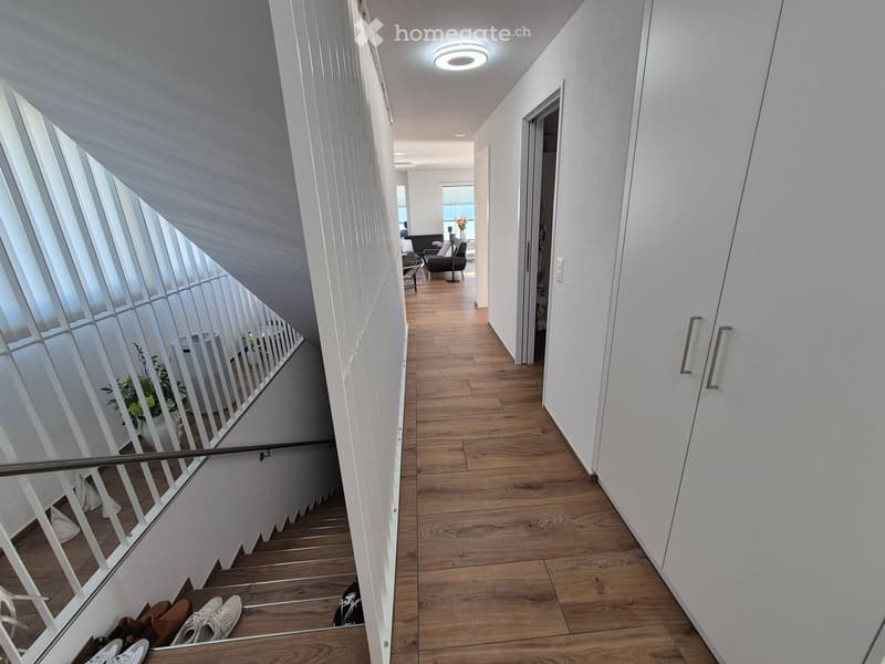 Modern hallway with wooden flooring, white walls, and a staircase leading to an upper level. The hallway features a sleek railing and a small seating area visible at the end.