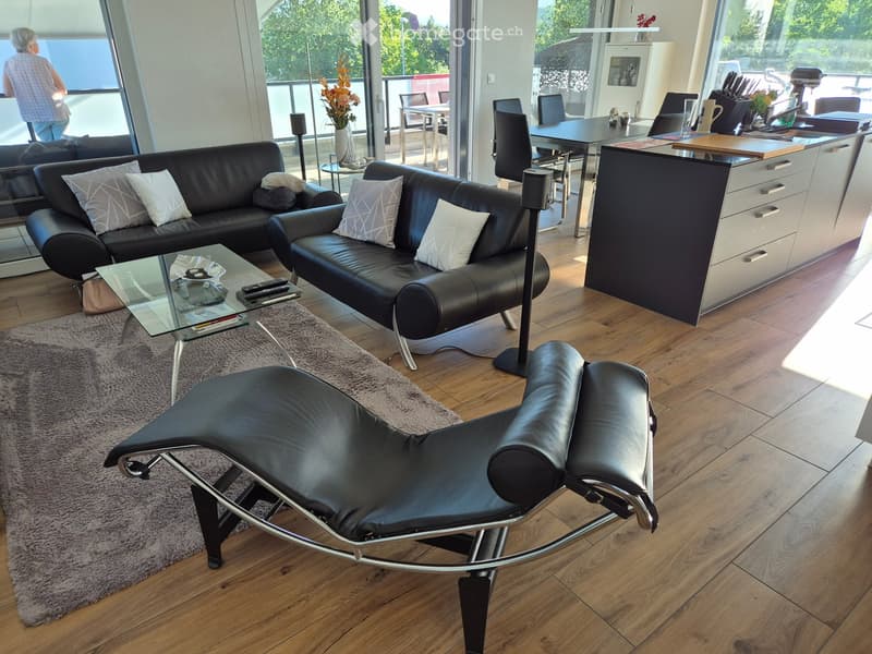 Modern living room with black leather sofas, a glass coffee table, a lounge chair, a kitchen island, and a dining area with chairs and a table.