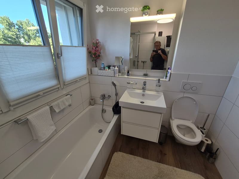 Modern bathroom with a bathtub, sink, toilet, large mirror, and window with blinds. Includes towels, a flower arrangement, and a small shelf with toiletries.