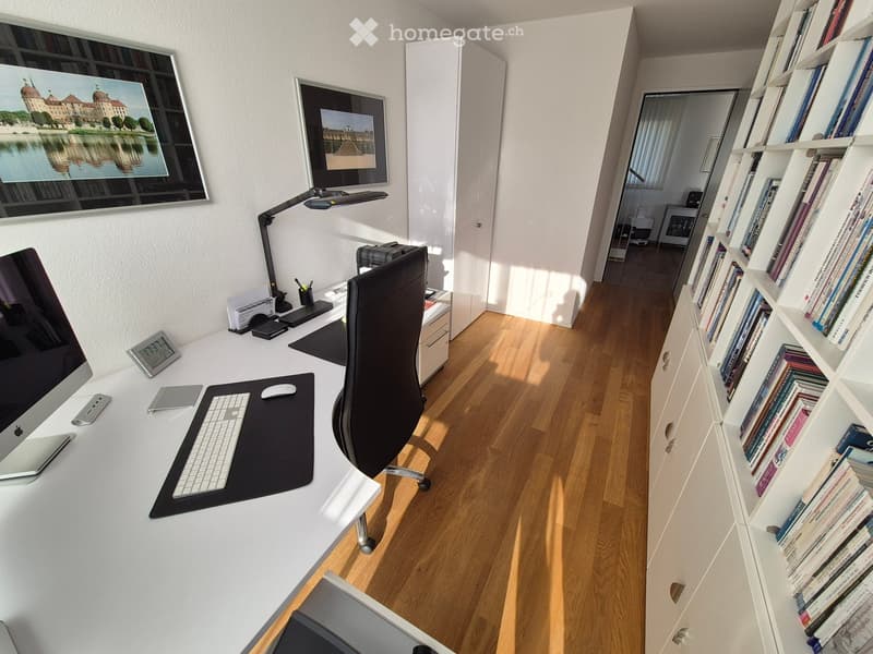Modern, well-lit office space with a white desk, ergonomic chair, computer setup, and a bookshelf filled with books. The room features wooden flooring and large windows allowing natural light.