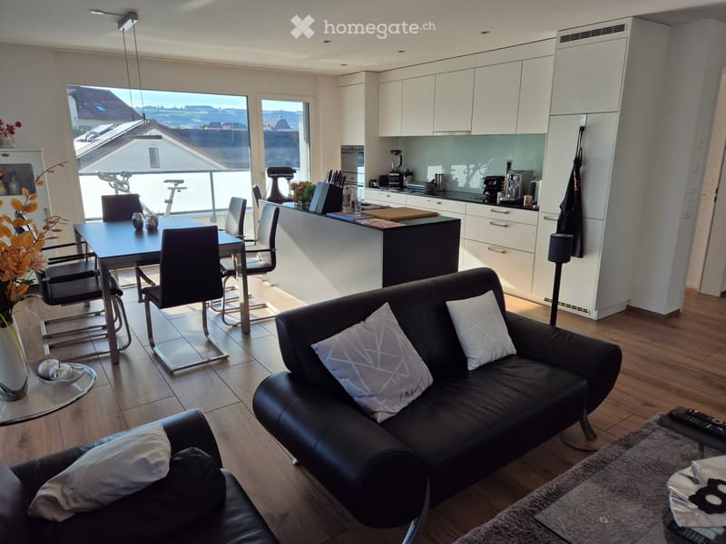 Modern open-plan living and dining area with a black leather sofa, dining table with chairs, kitchen area with white cabinets, and a view of the city from large windows.