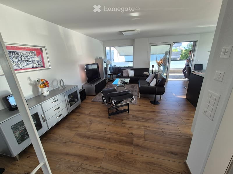 Modern living room with wooden flooring, large windows, and a balcony view. Features a black sofa, coffee table, armchair, TV, and decorative cabinet.