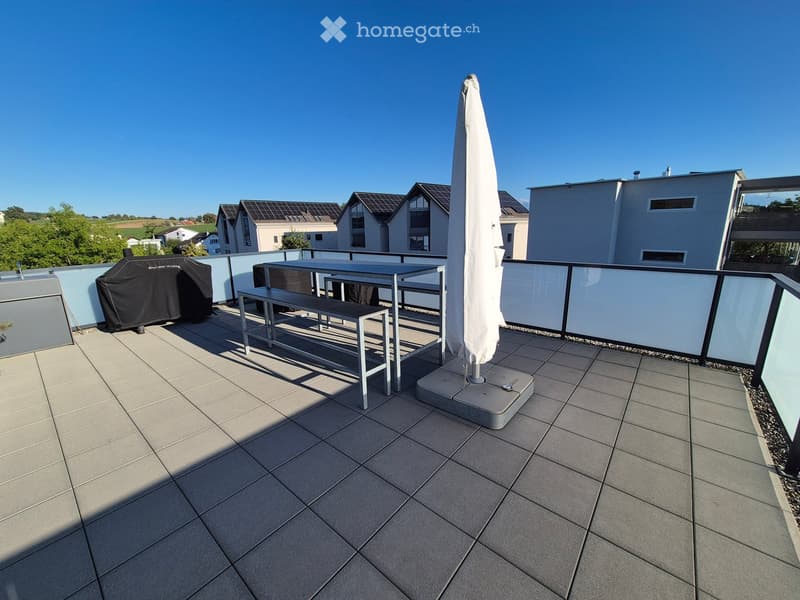 Balcony with tiled floor, metal table, bench, and umbrella, overlooking a residential area with modern houses and greenery.