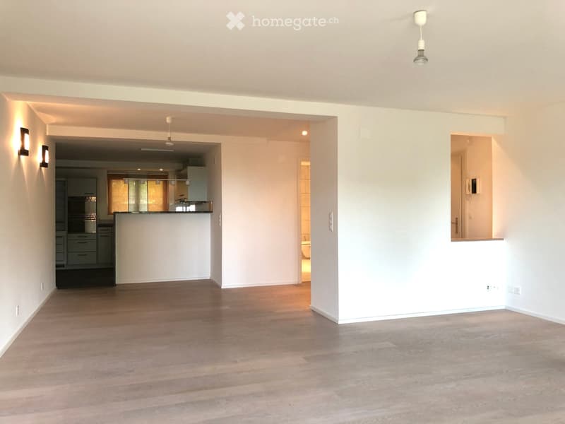 Empty room with light wooden flooring and white walls, featuring a modern pendant light fixture and an open doorway leading to a kitchen area.