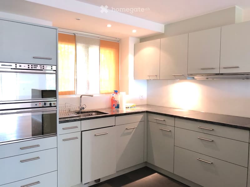Modern kitchen with white cabinets, stainless steel oven, sink, and countertop. Features a window with orange blinds and cleaning supplies on the counter.