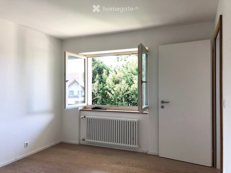 Empty room with light-colored walls, wooden flooring, a radiator, and a large window with a view of greenery and a neighboring house.