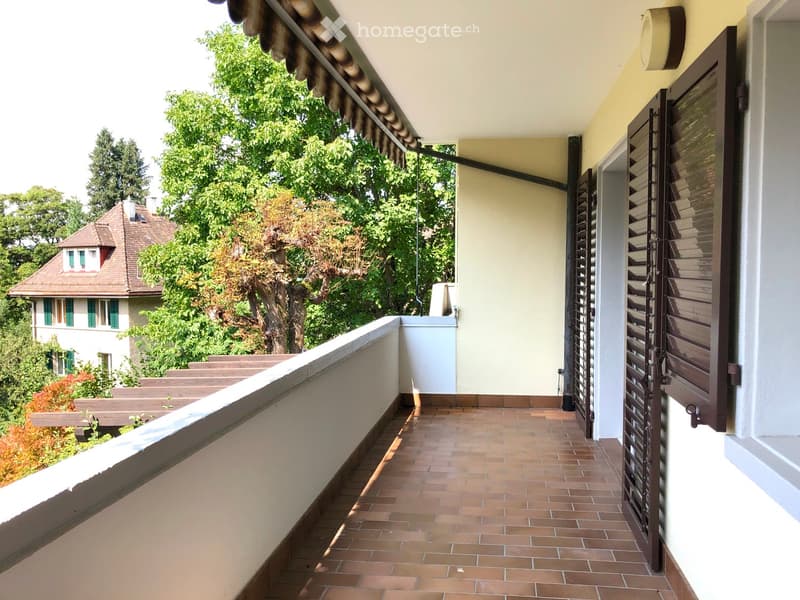 Balcony with tiled floor, wooden railing, and wooden shutters on the adjacent wall. The view includes a neighboring house with a red-tiled roof and green trees.