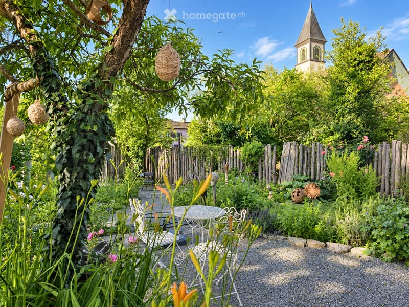 A lush garden with a small seating area featuring a white metal table and chairs, surrounded by greenery, flowers, and a wooden fence. A church tower is visible in the background.