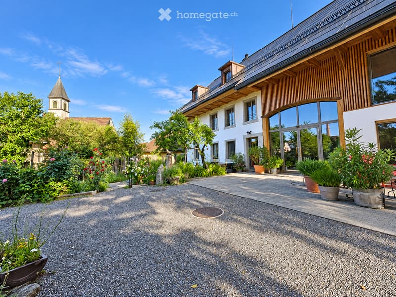 Garden area with gravel pathway, potted plants, and a modern house with large windows and wooden accents in the background.
