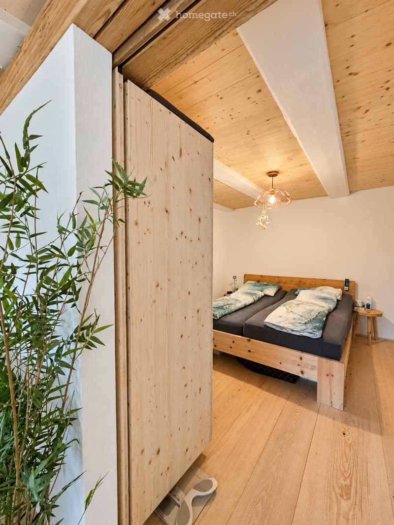Cozy bedroom with wooden bed, nightstand, and a hanging light fixture. The room features light wooden flooring and white walls, with a small potted plant near the entrance.