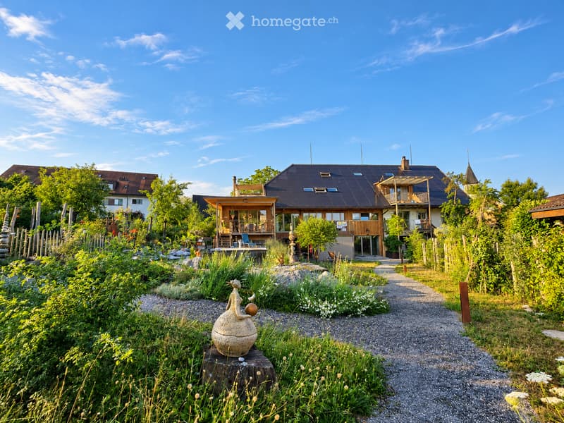 A modern house surrounded by a lush garden with pathways, sculptures, and outdoor seating areas.