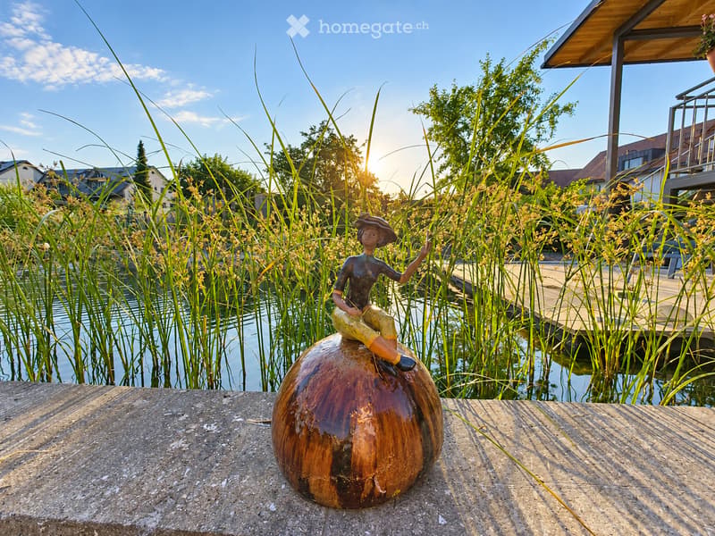 Garden area with a wooden deck, a small pond surrounded by tall reeds, and a decorative sculpture of a figure sitting on a wooden sphere.