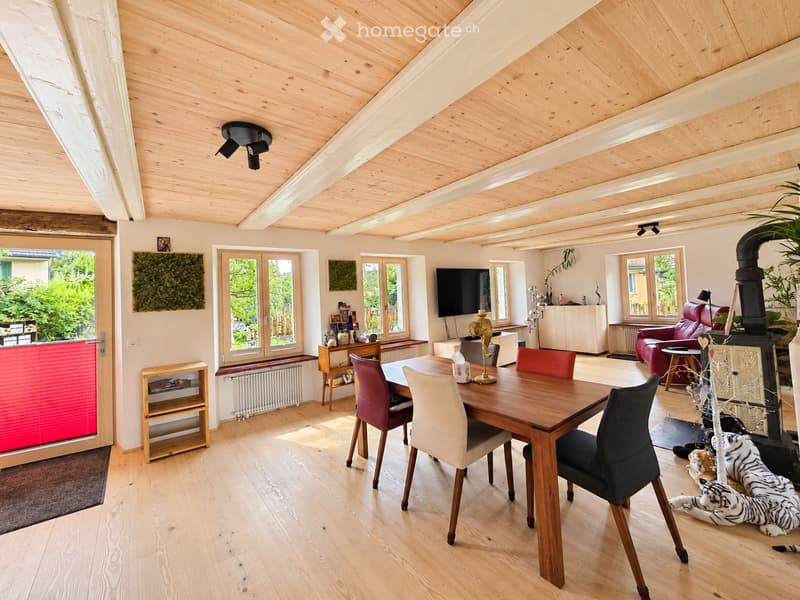 Bright, open-plan living and dining area with wooden floors, exposed wooden ceiling beams, large windows, and a fireplace. Features a dining table with chairs, a red leather sofa, a television, and decorative items.