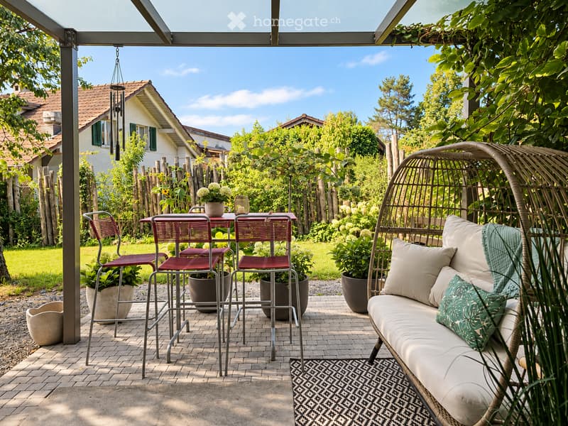 A well-maintained garden with a patio area featuring a wicker hanging egg chair, a small table with chairs, and potted plants. The background shows a house with a tiled roof and a wind chime hanging from the pergola.
