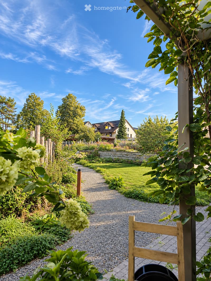 A well-maintained garden with a gravel pathway, lush greenery, and a stone wall. The garden features various plants, shrubs, and trees, with a wooden chair visible on the right side.