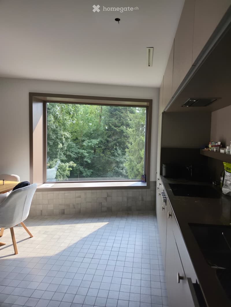 Modern kitchen with white tiled floor, large window overlooking greenery, white cabinetry, sink, and a small dining area with a table and chairs.