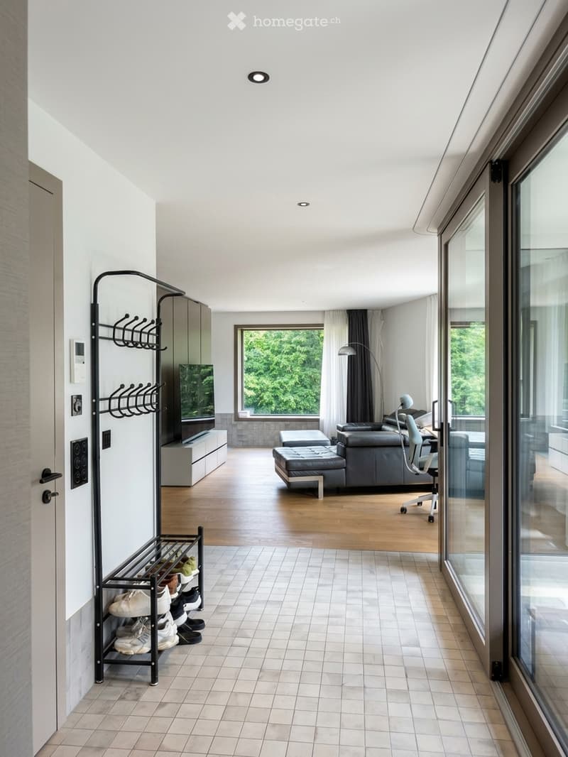 Modern living room with large windows, grey sofa, ottoman, chair, table, screen, lamp, and shoe rack.