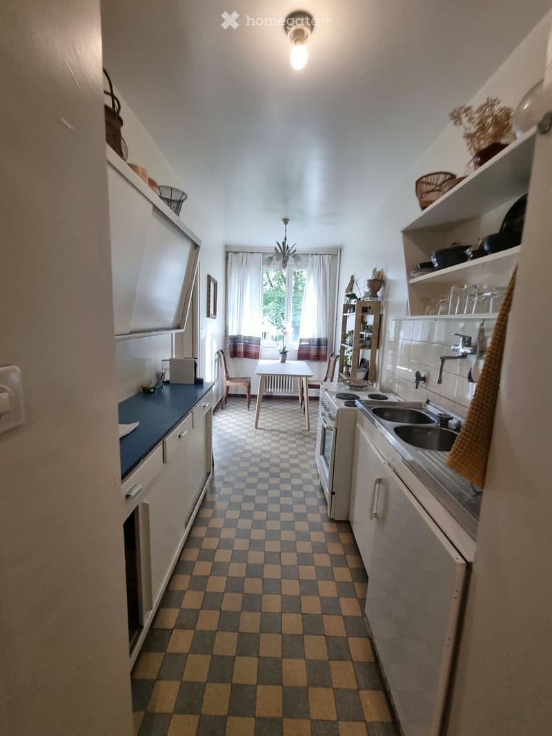 A small kitchen with checkered flooring, white cabinets, a sink, an oven, a small table with chairs, and shelves with various items.