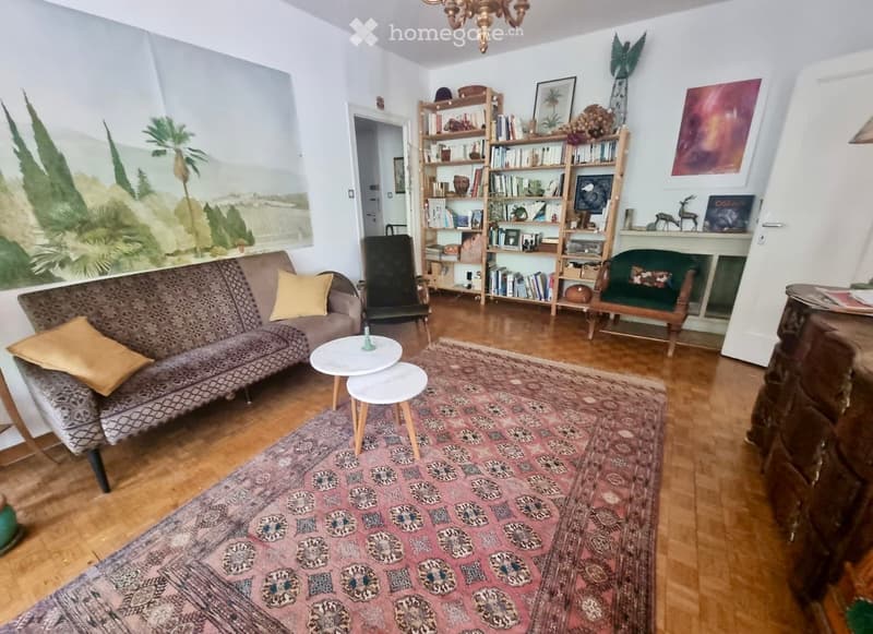 Living room with a patterned sofa, wooden floor, colorful rug, bookshelf, and small round tables.
