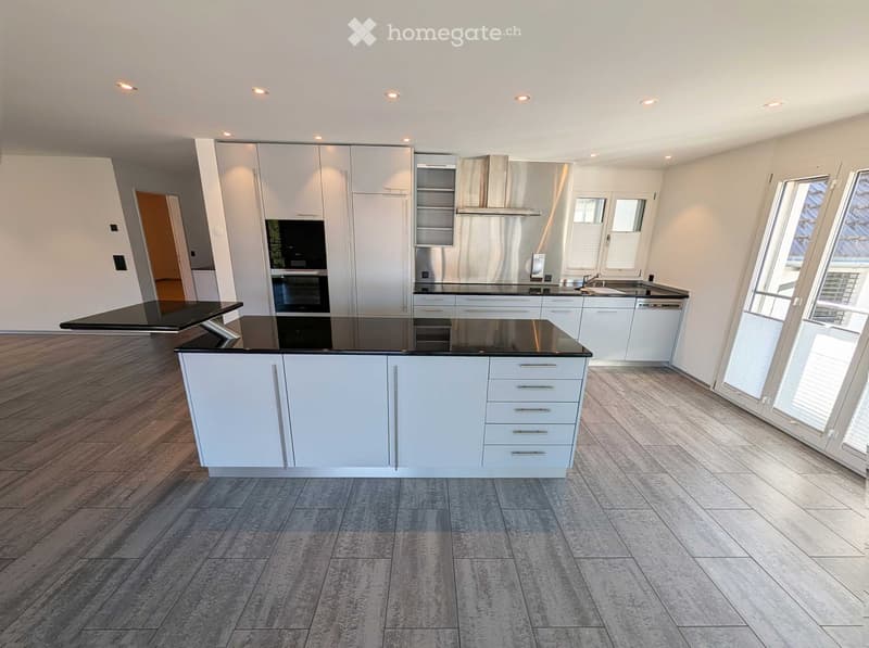 Modern kitchen with white cabinets, black countertops, and stainless steel appliances, including an oven and dishwasher. Large windows and recessed lighting.