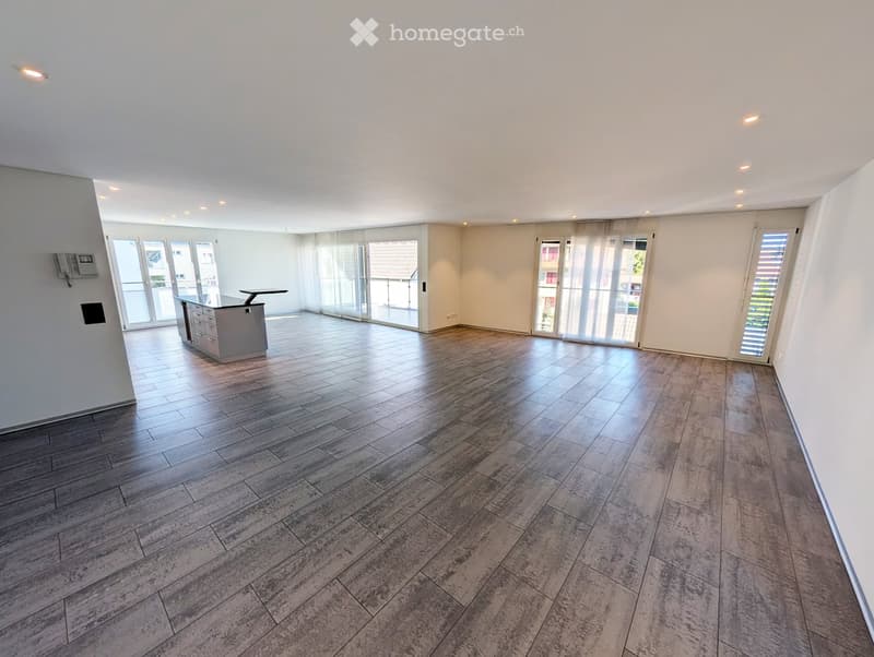 Empty room with wooden flooring, white walls, large windows, and a small kitchen area with cabinets and a countertop.