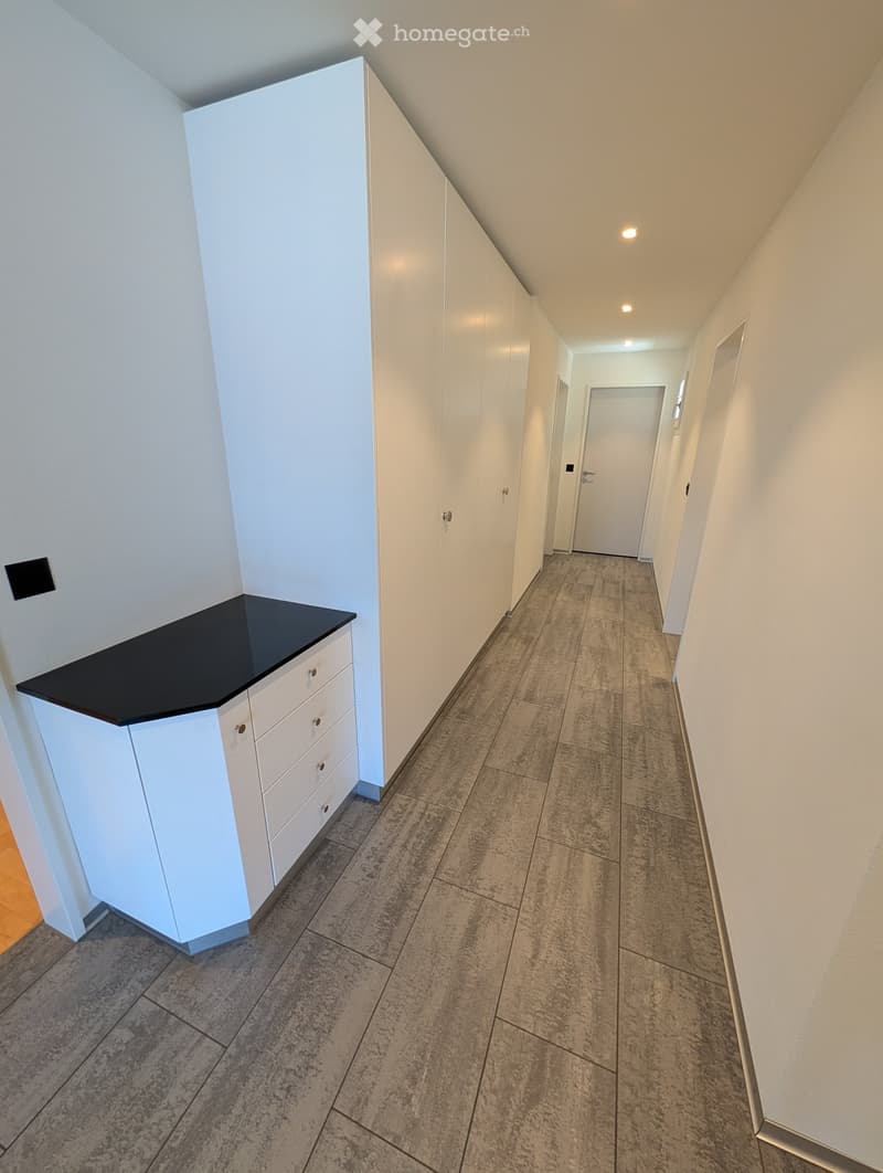 Clean, modern hallway with white walls, tiled floor, built-in white storage units, and a small white cabinet with a black countertop on the left side.