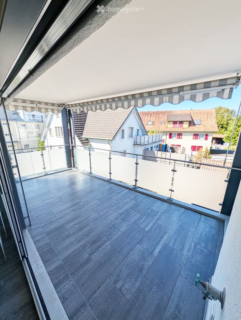 Balcony with tiled floor, glass railing, and striped awning, overlooking neighboring houses.