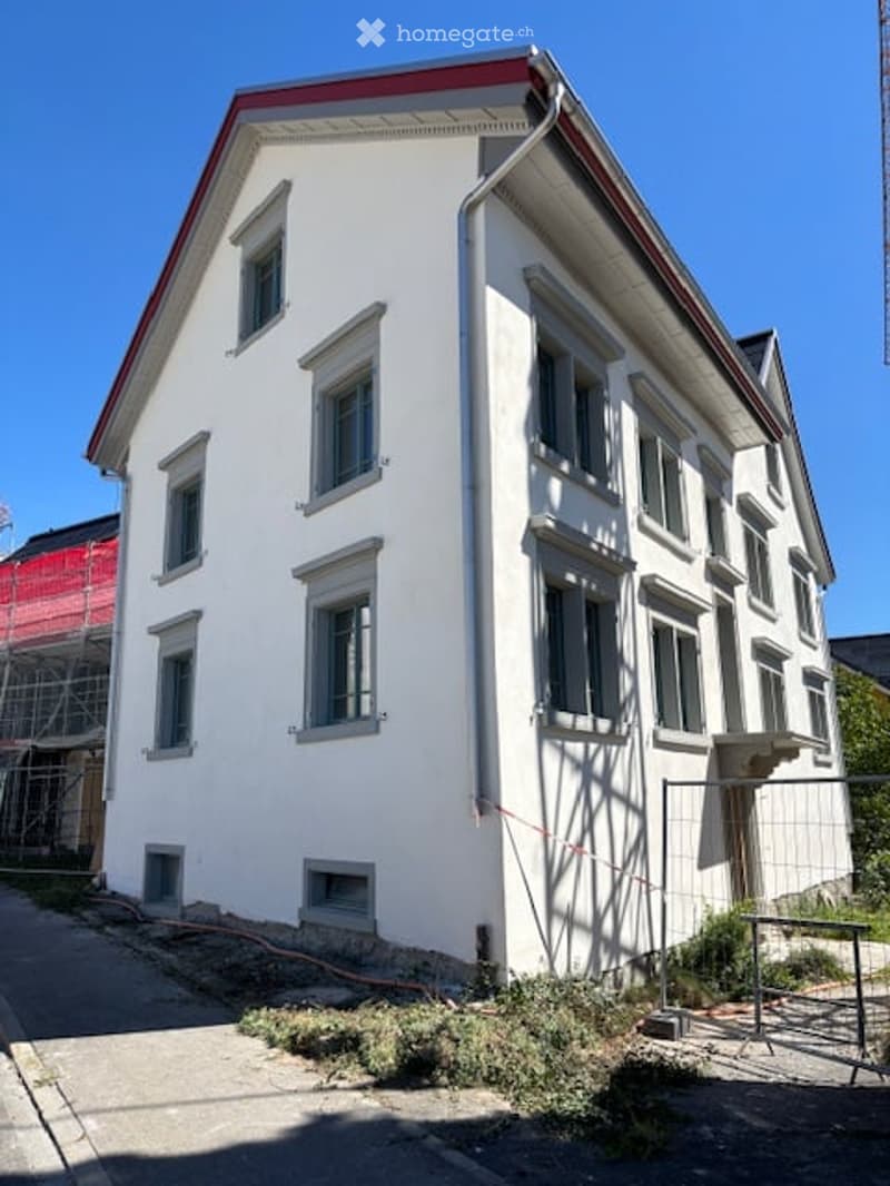 A two-story white building with multiple windows and a red roof, situated on a street with some construction barriers and greenery nearby.