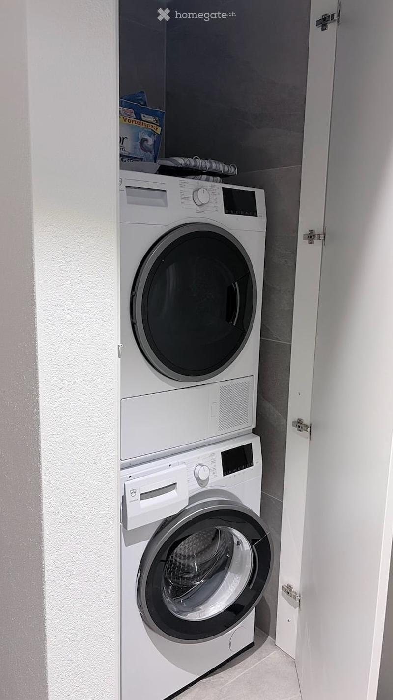 A compact laundry area featuring a stacked washer and dryer unit with a control panel, situated in a small, well-lit space.