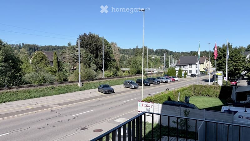 Street view showing a road with parked cars, a railway track, and residential buildings in the background.