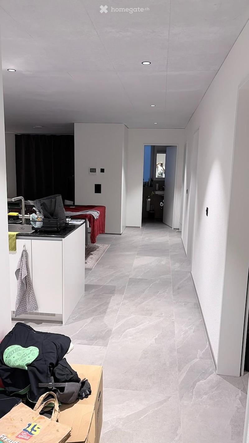 A modern hallway with a polished marble floor, white walls, and recessed lighting. Visible items include a kitchen area with a counter, sink, and cabinet, as well as a backpack, handbag, and laptop on a table.