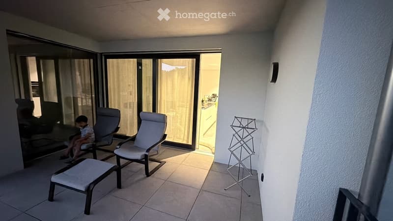 Balcony with tiled floor, two lounge chairs, a small table, and a metal rack. Large glass sliding doors open to an interior space.