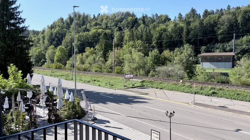 Balcony overlooking a street lined with closed umbrellas and outdoor seating, with a railway track and lush green forest in the background.