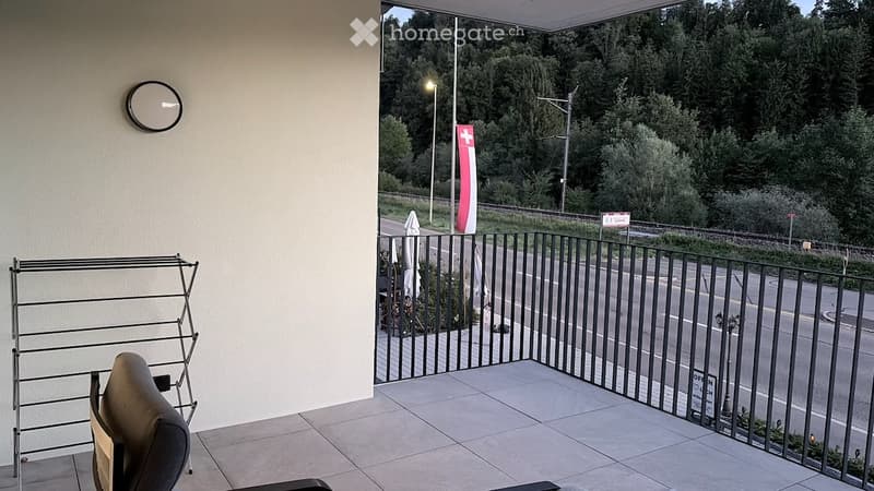 Balcony with tiled floor, black metal railing, view of road and greenery, umbrella, chair, and mirror on wall.