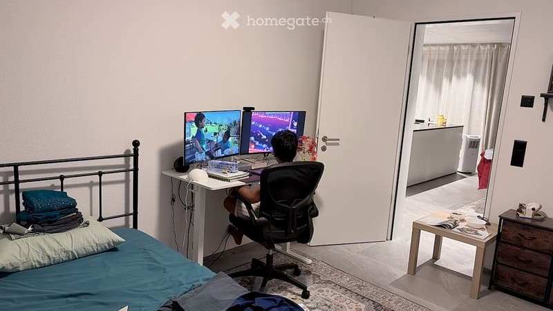 A modern bedroom with a bed, desk, and chair. The desk has a computer setup with two monitors, and there is a small table with a lamp and other items. The room features a carpeted floor and a partially open door leading to another room.
