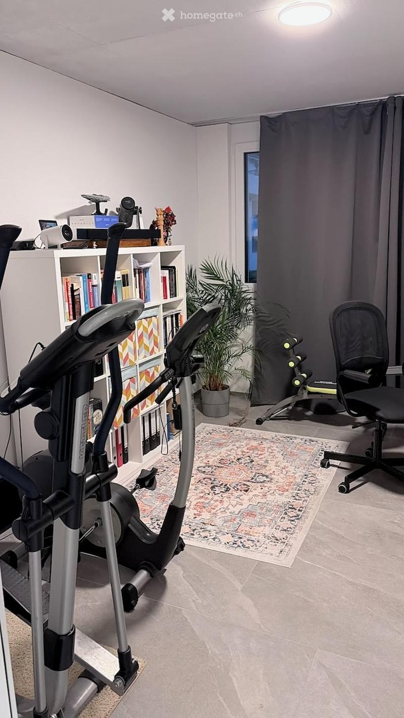 A well-lit room with a tiled floor, featuring an exercise bike, a bookshelf filled with books, a potted plant, and a comfortable chair.