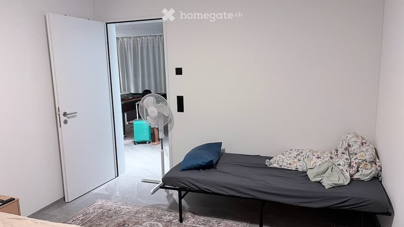 A small bedroom with a single bed, a standing fan, and an open door leading to another room with a desk and chair.