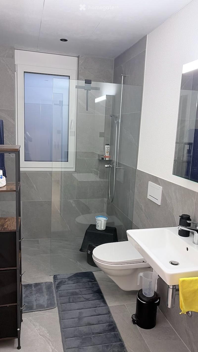 Modern bathroom with a glass-enclosed shower, toilet, sink, and mirror. Includes a small black stool with a cup, a yellow towel, and a gray bath mat on the floor.