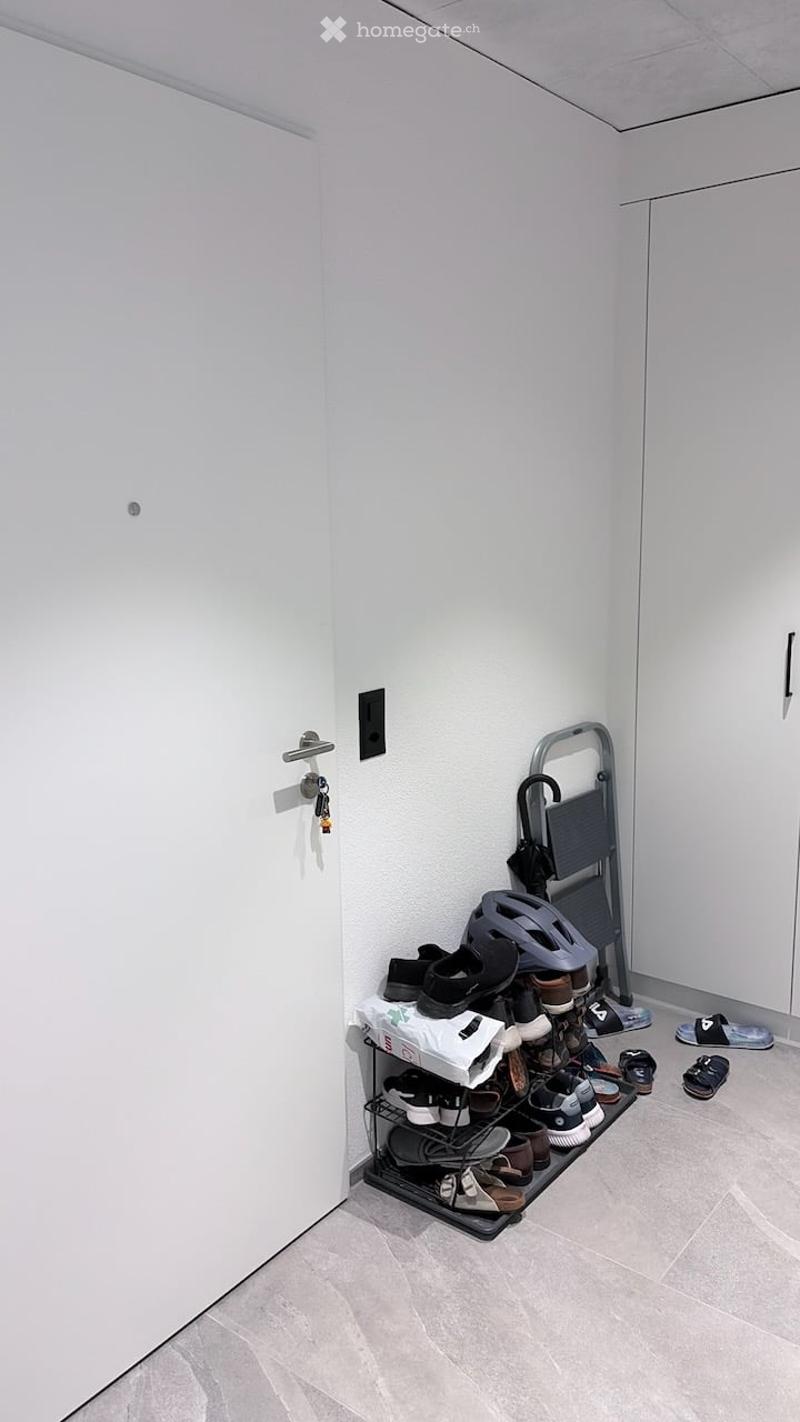 A narrow hallway with a shoe rack holding multiple pairs of shoes, a helmet, and a small ladder. The walls are white, and the floor is tiled.